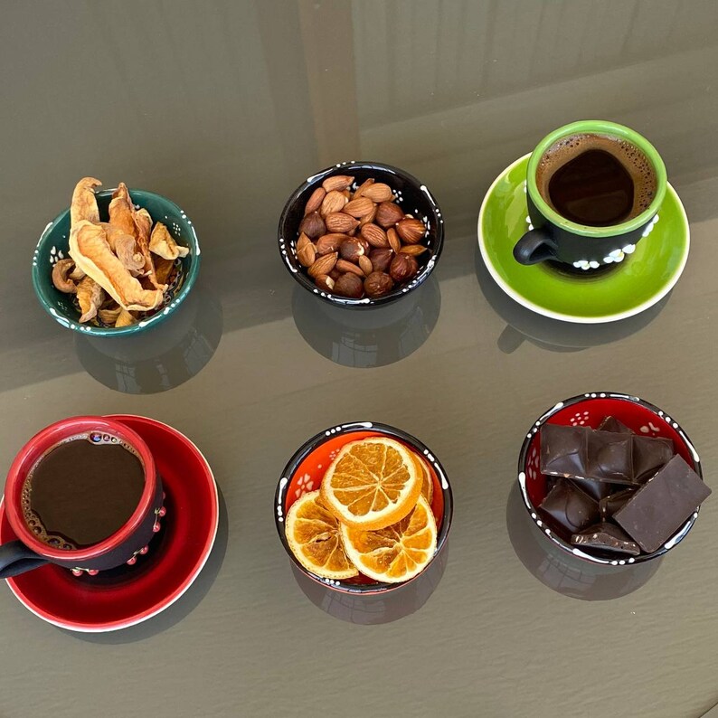 May include: Six small bowls with different snacks and two cups of coffee. The bowls are decorated with black and white patterns and have a red, green, or orange rim. The snacks include dried fruit, nuts, orange slices, and chocolate squares.