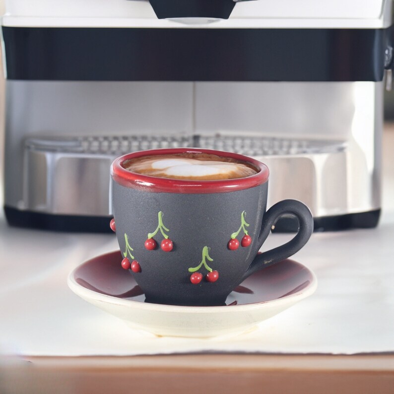 May include: A black ceramic coffee cup with a red rim and cherry designs, filled with a cappuccino with a heart-shaped design in the foam, sitting on a red saucer.