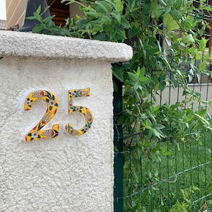 May include: A white ceramic house number with a yellow background and colorful floral designs. The number is 25.