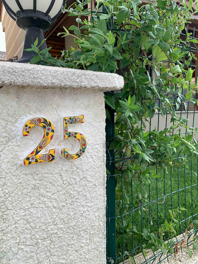 May include: A white ceramic house number with a yellow background and colorful floral designs. The number is 25.