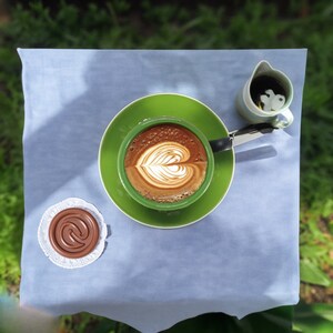May include: A green saucer with a white coffee cup with a heart-shaped design in the coffee. The coffee cup is on a blue tablecloth with a small chocolate-colored candy on a white doily.