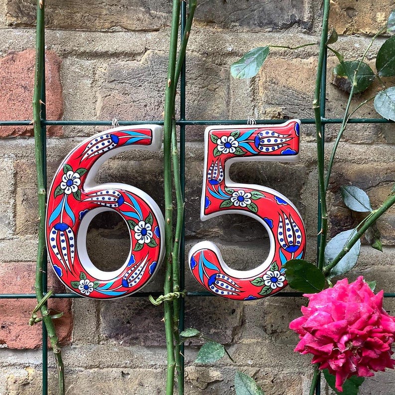 May include: Two ceramic house numbers, 6 and 5, painted in a red, white, and blue floral pattern. The numbers are attached to a green metal fence.
