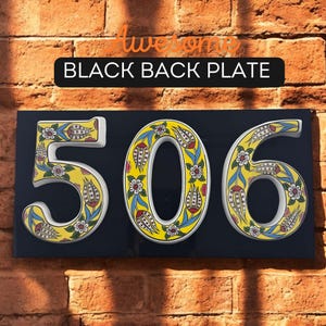 May include: A black ceramic house number plaque with white, yellow, blue, and red floral designs. The numbers 506 are displayed on the plaque.