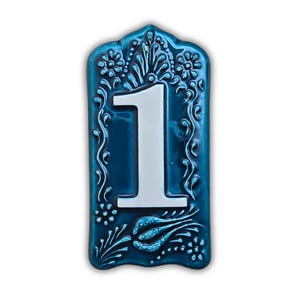 Pode incluir: Número de casa de cerâmica azul turquesa com o número um branco. O número está centrado na placa retangular. A placa tem um design decorativo de flores e videiras. As bordas da placa são recortadas.
