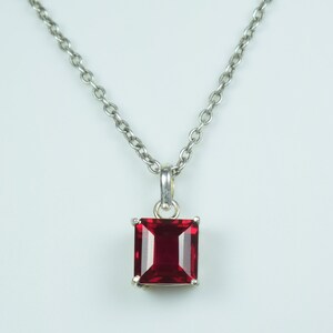 May include: A silver necklace with a rectangular, deep red gemstone pendant. The pendant is set in a silver frame and hangs from a delicate silver chain. The gemstone has a faceted surface.