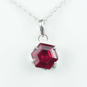 May include: A silver necklace featuring a hexagonal, faceted, deep red gemstone pendant. The pendant is held in place by four silver prongs and hangs from a delicate silver chain. The gemstone has a brilliant, reflective surface.