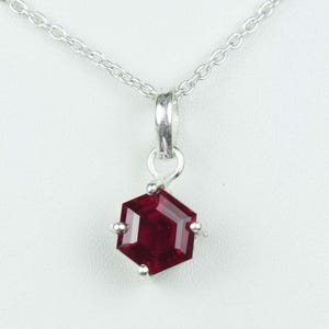 May include: A silver necklace featuring a hexagonal, deep red pendant. The faceted pendant hangs from a delicate chain. The necklace is shown against a white backdrop.