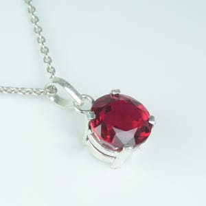 May include: A silver necklace featuring a large, oval-cut, deep red gemstone pendant. The faceted gemstone reflects light. The delicate chain supports the pendant, which is set in a silver metal setting. The background is a soft, neutral colour.