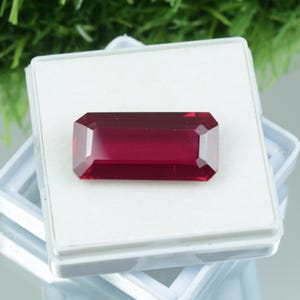 Peut inclure: Une pierre précieuse rouge foncé, rectangulaire et facettée, peut-être un rubis, présentée dans un boîtier en plastique blanc. La gemme a une surface polie et une taille symétrique, reflétant la lumière. Le boîtier est ouvert, révélant la couleur vibrante de la gemme.