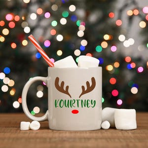 Może przedstawiać: Biały kubek ceramiczny z brązowym wzorem renifera i imieniem "Kourtney" w zielonych literach. Kubek ma czerwony nos i jest wypełniony piankami. Słomka w czerwono-białe paski wystaje z kubka.