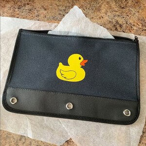 May include: A black pencil case with a yellow rubber ducky design. The case has three grommets on the bottom and a zipper closure.