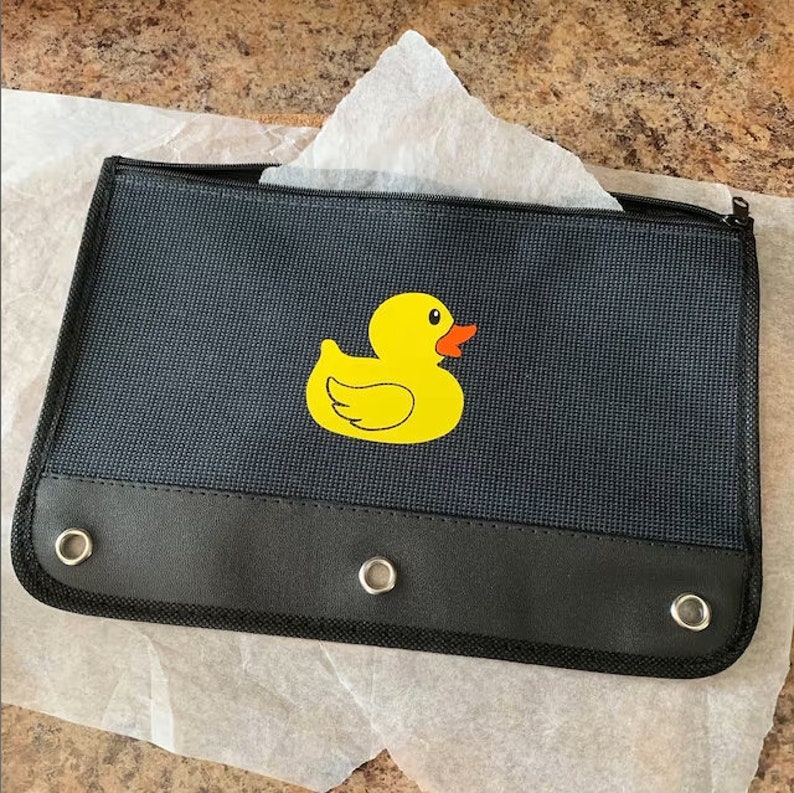 May include: A black pencil case with a yellow rubber ducky design. The case has three grommets on the bottom and a zipper closure.