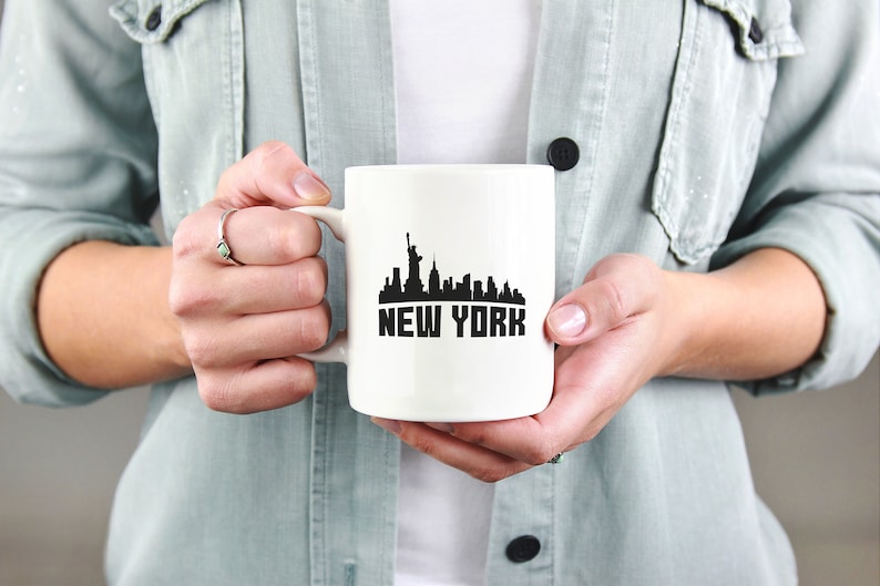 May include: White ceramic mug with a black silhouette of the New York City skyline and the text "NEW YORK" printed on the front.