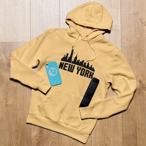May include: A mustard yellow hoodie with a black New York City skyline graphic and the words "NEW YORK" printed on the front. A blue phone case and a black remote control are lying on the hoodie.