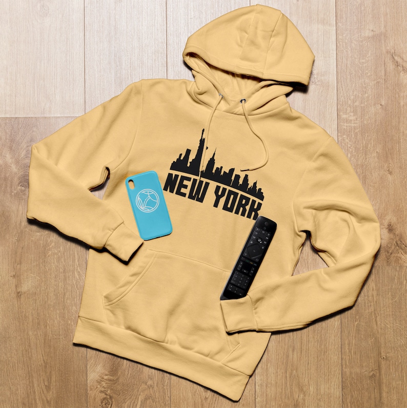May include: A mustard yellow hoodie with a black New York City skyline graphic and the words "NEW YORK" printed on the front. A blue phone case and a black remote control are lying on the hoodie.