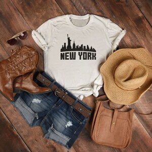 May include: A white t-shirt with a black silhouette of the New York City skyline and the text "NEW YORK". The shirt is laid out on a wooden surface with a pair of brown cowboy boots, a denim shorts, a brown leather bag, and a straw hat.