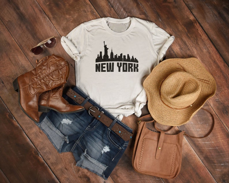 May include: A white t-shirt with a black silhouette of the New York City skyline and the text "NEW YORK". The shirt is laid out on a wooden surface with a pair of brown cowboy boots, a denim shorts, a brown leather bag, and a straw hat.