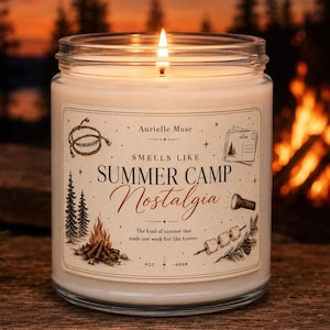 May include: A clear glass jar candle with a lit wick and white wax. The label reads Aurielle Muse, Smells Like Summer Camp Nostalgia, and includes illustrations of a campfire, pine trees, a postcard, and marshmallows on a stick. It is 9oz.