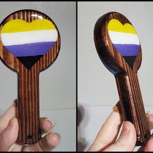 Peut inclure: Une décoration en bois en forme de cœur peinte avec les couleurs du drapeau de la fierté non binaire : jaune, blanc, violet et noir. La décoration a une poignée en bois brun.