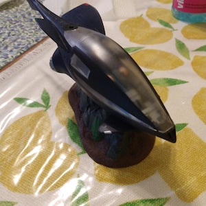 May include: A dark grey and black spaceship model with a silver-coloured canopy. The model is sitting on a brown base with green accents.