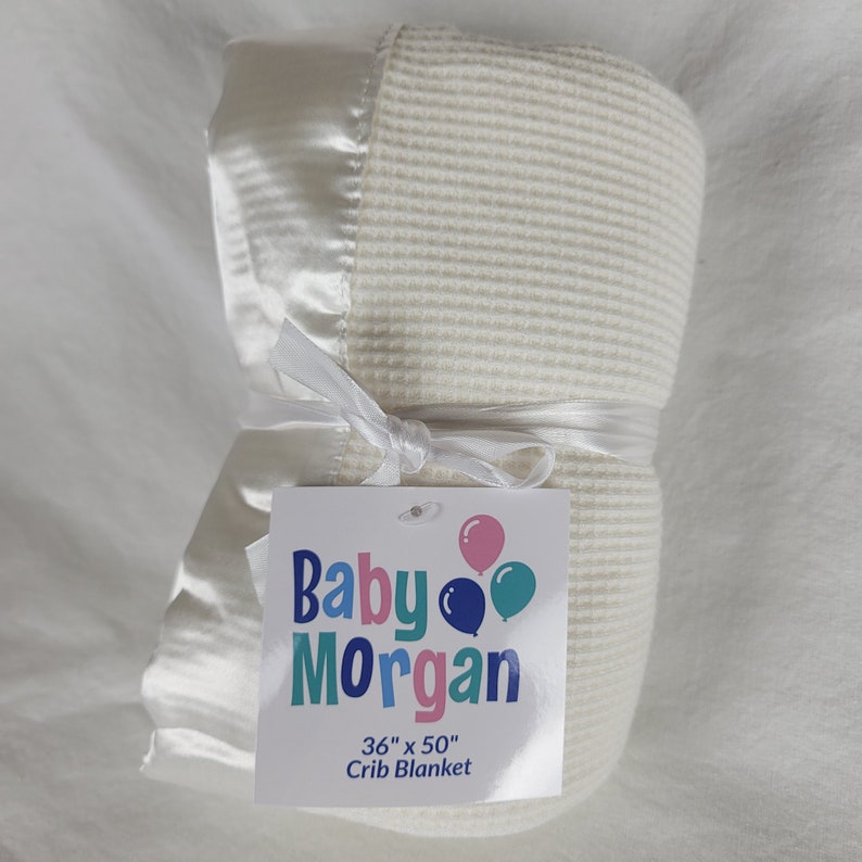 K&ouml;nnte beinhalten: Eine wei&szlig;e Babydecke mit Satinbord&uuml;re, aufgerollt und mit einem wei&szlig;en Band gebunden. Die Decke hat ein Etikett mit der Aufschrift "Baby Morgan 36" x 50" Babydecke".