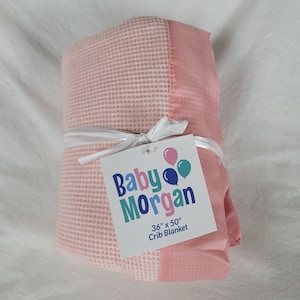 K&ouml;nnte beinhalten: Eine rosa und wei&szlig;e Waffelstruktur-Babydecke mit rosa Satin-Bord&uuml;re. Die Decke ist mit einem wei&szlig;en Band gebunden und hat ein Etikett mit der Aufschrift "Baby Morgan 36" x 50" Crib Blanket".