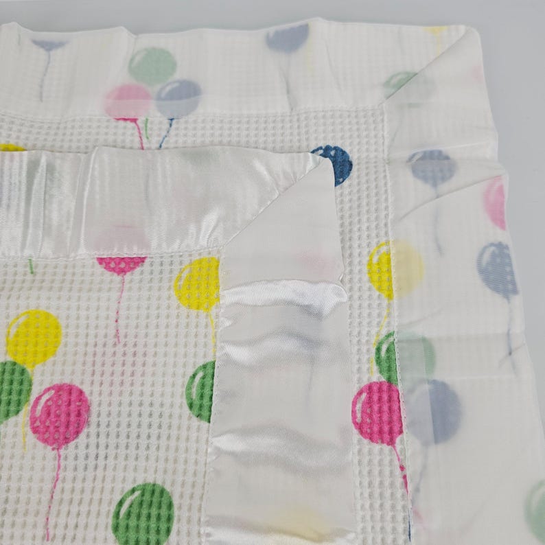 May include: A white waffle weave baby blanket with a satin trim and a colorful balloon print. The balloons are pink, yellow, green, and blue.