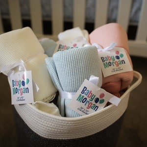 K&ouml;nnte beinhalten: Ein Korb gef&uuml;llt mit weichen, Waffel-Web-Babydecken in verschiedenen Farben, darunter Wei&szlig;, Hellblau und Rosa. Jede Decke hat ein Etikett mit der Aufschrift "Baby Morgan 34" x 50" Crib Blanket".