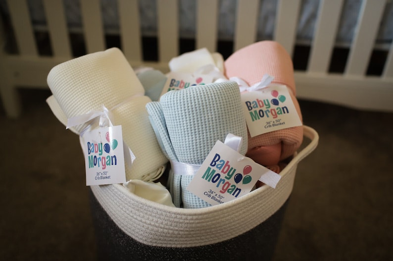 K&ouml;nnte beinhalten: Ein Korb gef&uuml;llt mit weichen, Waffel-Web-Babydecken in verschiedenen Farben, darunter Wei&szlig;, Hellblau und Rosa. Jede Decke hat ein Etikett mit der Aufschrift "Baby Morgan 34" x 50" Crib Blanket".