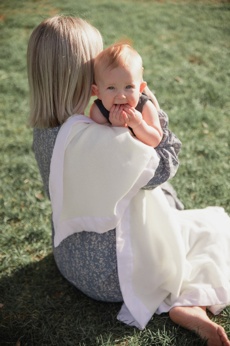 K&ouml;nnte beinhalten: Eine Frau in einem blauen Blumenkleid h&auml;lt ein Baby, das in eine wei&szlig;e Strickdecke gewickelt ist. Das Baby schaut in die Kamera und hat eine Hand im Mund.