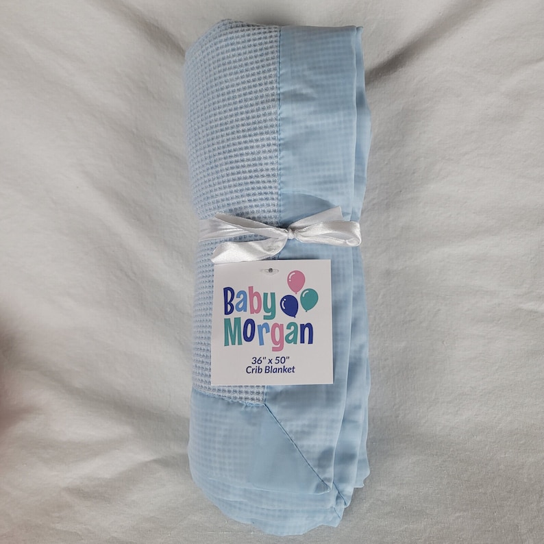 K&ouml;nnte beinhalten: Eine hellblaue Babydecke mit wei&szlig;em Satinrand. Die Decke ist aufgerollt und mit einem wei&szlig;en Band gebunden. Ein wei&szlig;es Etikett mit dem Text "Baby Morgan 36" x 50" Crib Blanket" ist am Band befestigt.