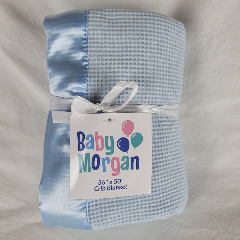 K&ouml;nnte beinhalten: Eine hellblaue Waffel-Decke f&uuml;r Babybetten mit Satinbesatz. Die Decke ist mit einem wei&szlig;en Band gebunden und hat ein Etikett mit der Aufschrift "Baby Morgan 36" x 50" Crib Blanket".