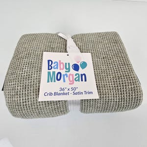 May include: A gray waffle weave crib blanket with a satin trim. The blanket is folded in half and has a tag attached that reads "Baby Morgan 36" x 50" Crib Blanket - Satin Trim".