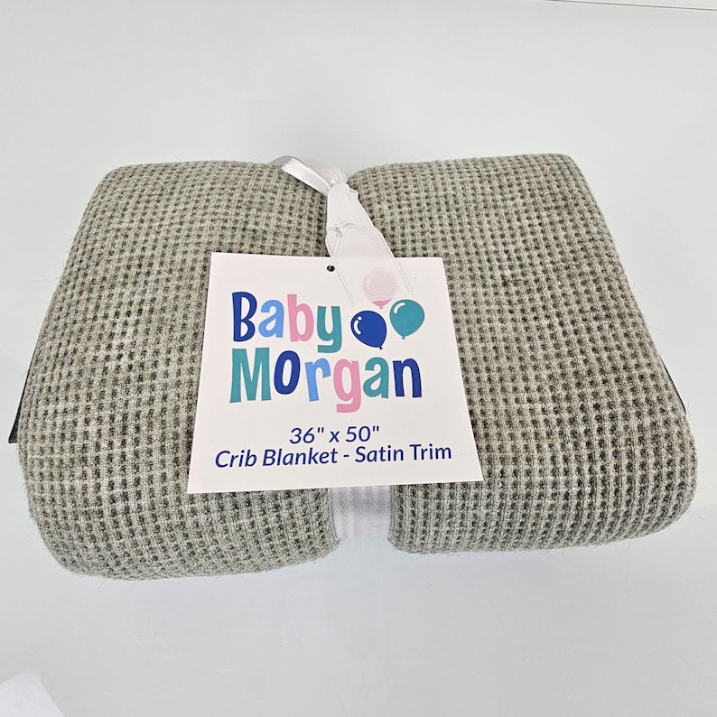 May include: A gray waffle weave crib blanket with a satin trim. The blanket is folded in half and has a tag attached that reads "Baby Morgan 36" x 50" Crib Blanket - Satin Trim".