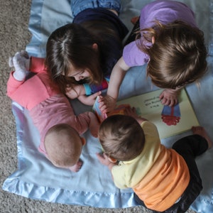 K&ouml;nnte beinhalten: Drei Kinder sitzen auf einer blauen Decke und lesen gemeinsam ein Buch. Das Buch ist ge&ouml;ffnet und zeigt eine bunte Illustration. Die Kinder l&auml;cheln alle und scheinen ihre Zeit zusammen zu genie&szlig;en.