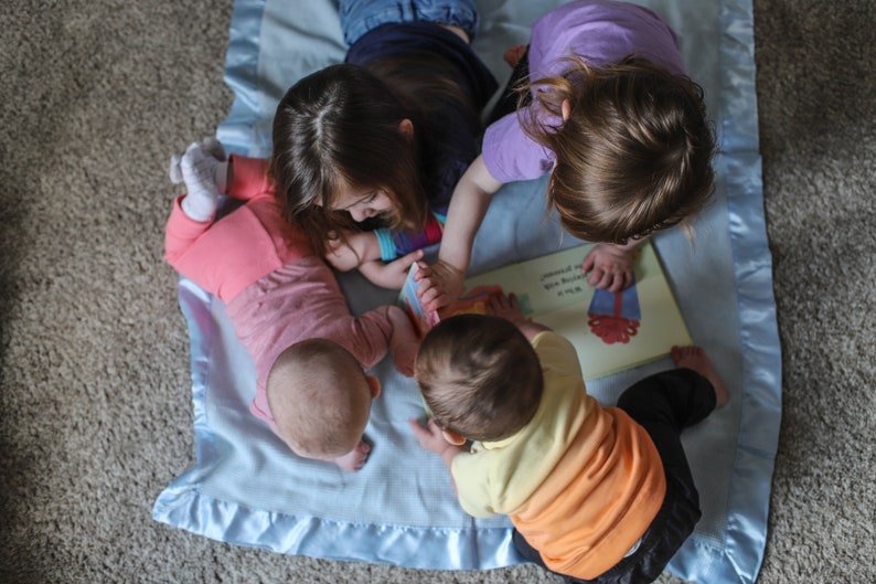 K&ouml;nnte beinhalten: Drei Kinder sitzen auf einer blauen Decke und lesen gemeinsam ein Buch. Das Buch ist ge&ouml;ffnet und zeigt eine bunte Illustration. Die Kinder l&auml;cheln alle und scheinen ihre Zeit zusammen zu genie&szlig;en.