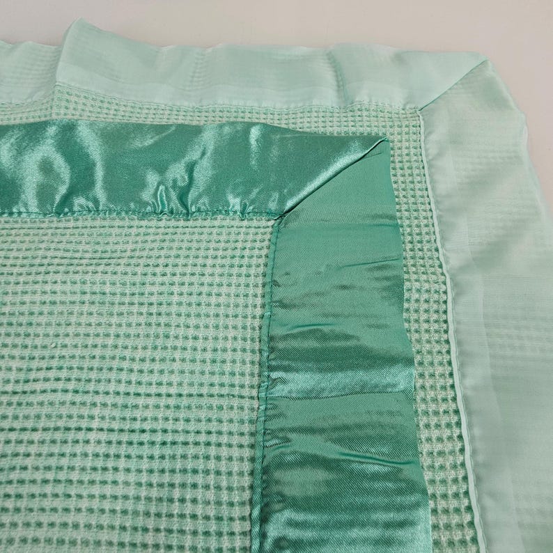May include: A soft, green waffle weave baby blanket with a satin trim. The blanket has a light green satin border and a darker green satin ribbon around the edge.