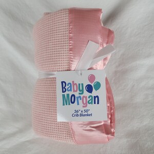 K&ouml;nnte beinhalten: Eine rosa und wei&szlig;e Waffel-Decke mit Satinbesatz. Die Decke ist aufgerollt und mit einem wei&szlig;en Band gebunden. Ein wei&szlig;es Etikett mit dem Text "Baby Morgan 36" x 50" Crib Blanket" ist an der Decke befestigt.