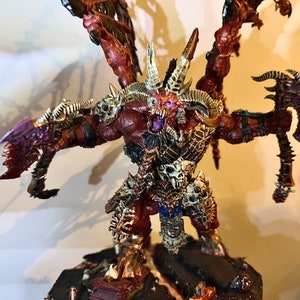 Puede incluir: Una miniatura pintada de rojo y dorado de un demonio con múltiples brazos y alas. El demonio sostiene un hacha grande en una mano y tiene una cara de calavera con ojos rojos brillantes.
