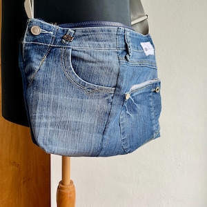 Könnte beinhalten: Blaue Jeans-Umhängetasche aus recycelten Jeans. Die Tasche hat einen Reißverschluss, Vordertaschen und einen Knopfverschluss. Sie hat einen dunkelblauen Riemen und ein weißes Etikett.