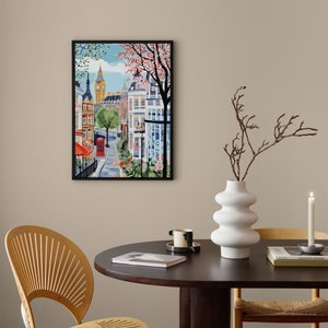 May include: A framed illustration of a street scene in London, England. The illustration features a red telephone booth, a tall building with a clock tower, and a street lined with colorful buildings. The illustration is painted in a bright, whimsical style.