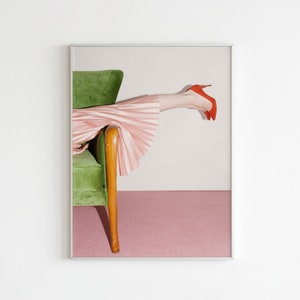 Puede incluir: Una pierna de mujer con una falda plisada rosa y tacones rojos descansa sobre un sillón de terciopelo verde. El sillón tiene patas de madera y está colocado sobre un fondo rosa claro.