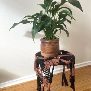 May include: A potted peace lily plant sits on a black and pink macrame stool with black metal legs.
