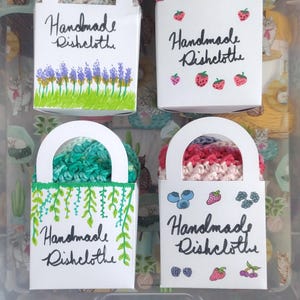 Könnte beinhalten: Vier weiße Boxen mit Griffen, jede mit einem handgefertigten Geschirrtuch. Die Boxen sind mit verschiedenen Blumen- und Beerenmustern verziert. Der Text "Handmade Dishcloth" ist auf jeder Box geschrieben.