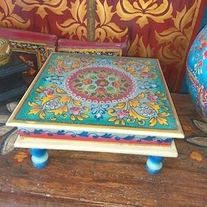 May include: A square wooden low table featuring a hand-painted top with a central floral medallion in shades of red, green, and yellow. The edges are decorated with blue leaf patterns, and the table is supported by four short, rounded blue legs.