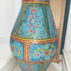 May include: A large, turquoise metal vase with hand-painted floral designs in red, orange, and pink. The vase has a cylindrical top and a wider, bulbous body, with a decorative ring handle. Yellow and white accents frame the floral patterns.