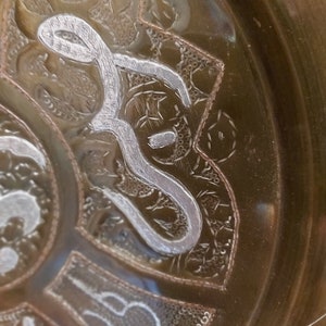 May include: Close-up of a decorative metal plate with intricate silver designs. The plate has a brown, metallic finish and features a repeating pattern of stylized floral motifs.