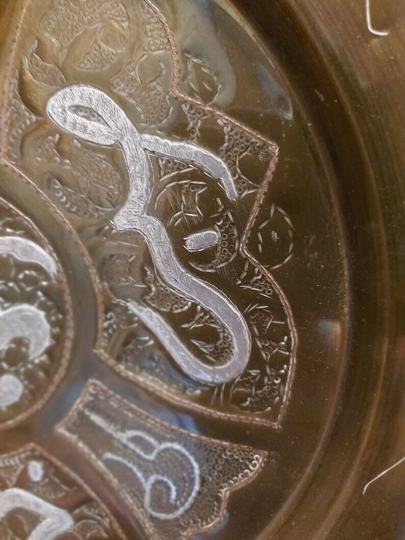 May include: Close-up of a decorative metal plate with intricate silver designs. The plate has a brown, metallic finish and features a repeating pattern of stylized floral motifs.