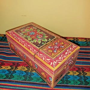 May include: A rectangular wooden box with a hinged lid, painted with floral designs in red, yellow, green, and blue. The box has a dark brown top with a floral pattern and a metal clasp.