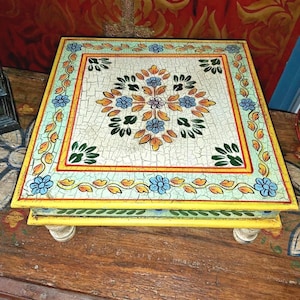 May include: A square wooden footed stool featuring a crackled white surface with a central floral motif in blue, orange, and green. The border displays a repeating vine pattern in yellow, green, and blue, with a bright yellow painted edge.